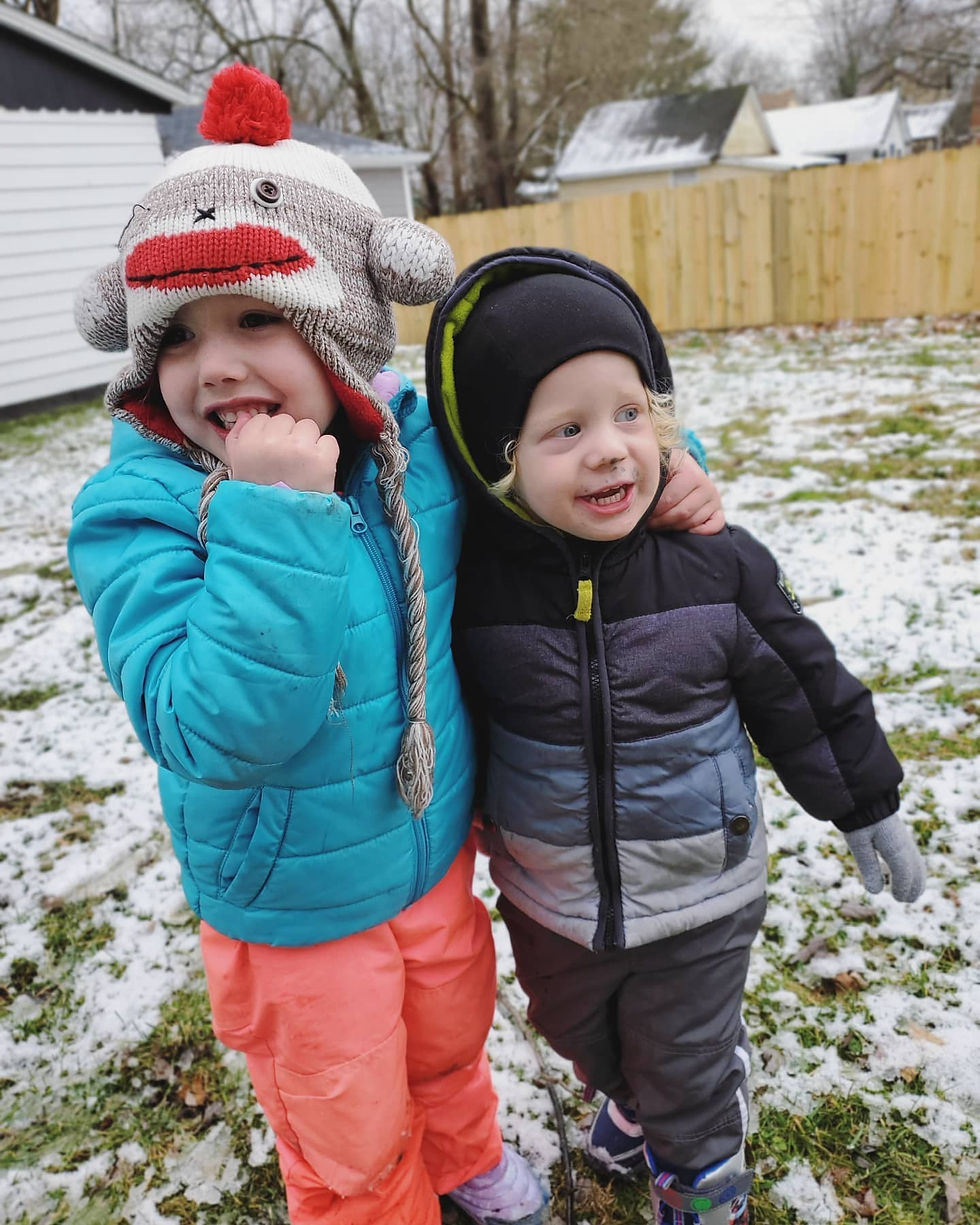 2 friends have arms around each other. they are in full snow gear. there is a dusting of snow on the ground. 