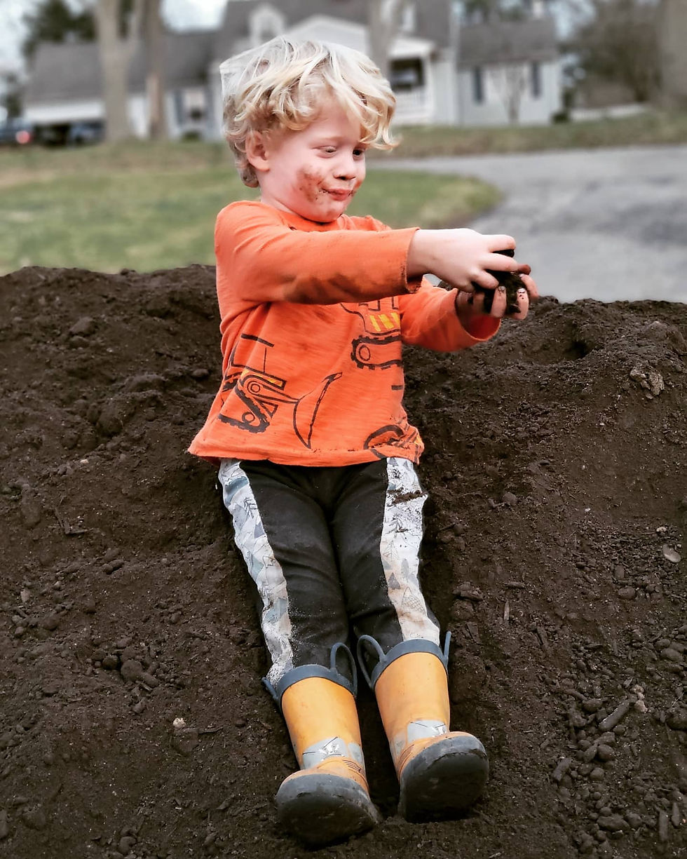 3 year old boy in orange shirt with yellow boots and black pants plays in dirt pile