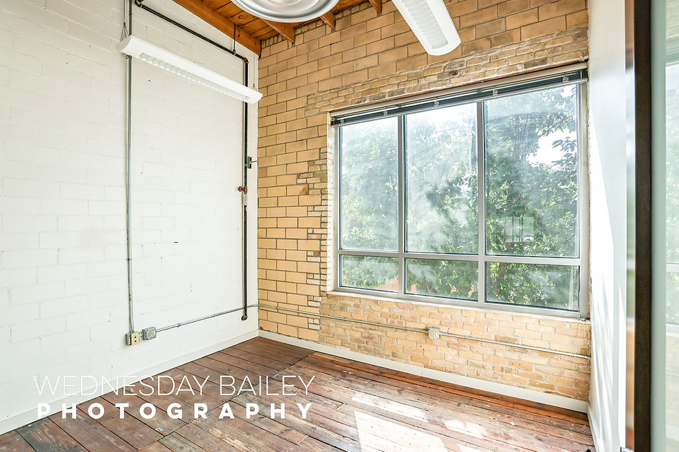 Sunlit room with brick walls, large window, and wooden floor. Pipes and light fixtures visible. Photo by Wednesday Bailey Photography.