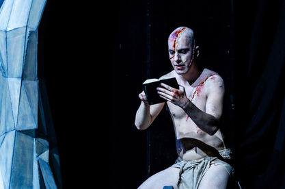 The shirtless, scarred creature sits next to a jagged tree, reading from a small black book.