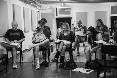 Cast members sit in a room reading music scores during a ZEST rehearsal for the musical RENT.
