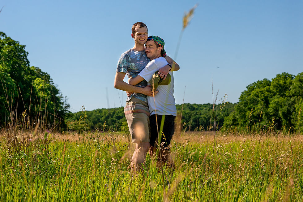 a photo of two young men standing in a meadow of tall grass hugging each other close. One young man has his eyes closed peacefully and his head rests on his partner's shoulder. The other young man is smiling widely.