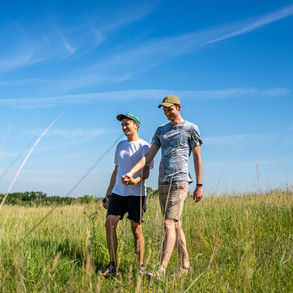 A photo of a gay couple holding hands and walking through a meadow tall grass with a bright blue sky behind.