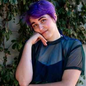 A photo of a feminine-presenting non binary person posing for the camera in front of a vine-covered wall. They have short bright purple hair and are wearing a sheer dark blue shirt over a dark spaghetti strap. They have their head tilted slightly to the left and are resting their cheek against their hand.