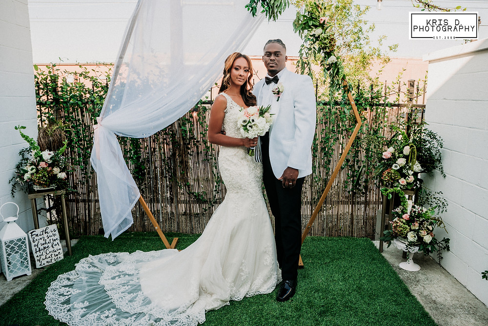 bride with lace train and groom in white tuxedo jacket posing in front of veiled and floral trellis