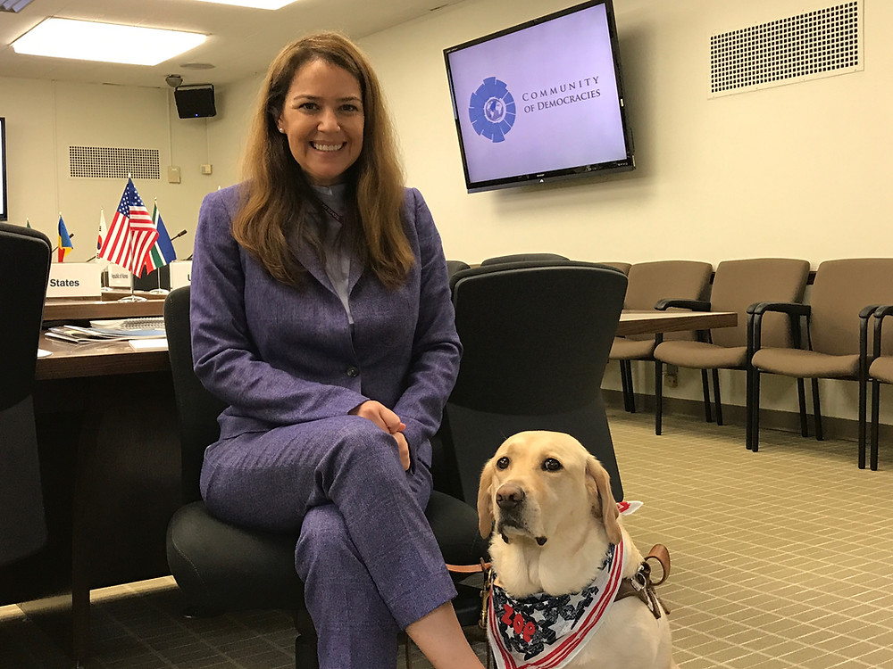PD: Kristin and Zoe are sitting in a conference room. Kristin is sitting on a chair, dressed in a formal blue-grey pantsuit, and smiling at the camera. Zoe is sitting on the ground at Kristin’s side, looking at the camera while wearing a USA themed bandana that reads “Zoe”. Behind them are several conference room chairs, a conference table, a few tabletop flags for various countries, and a TV screen that says “COMMUNITY OF DEMOCRACIES.”