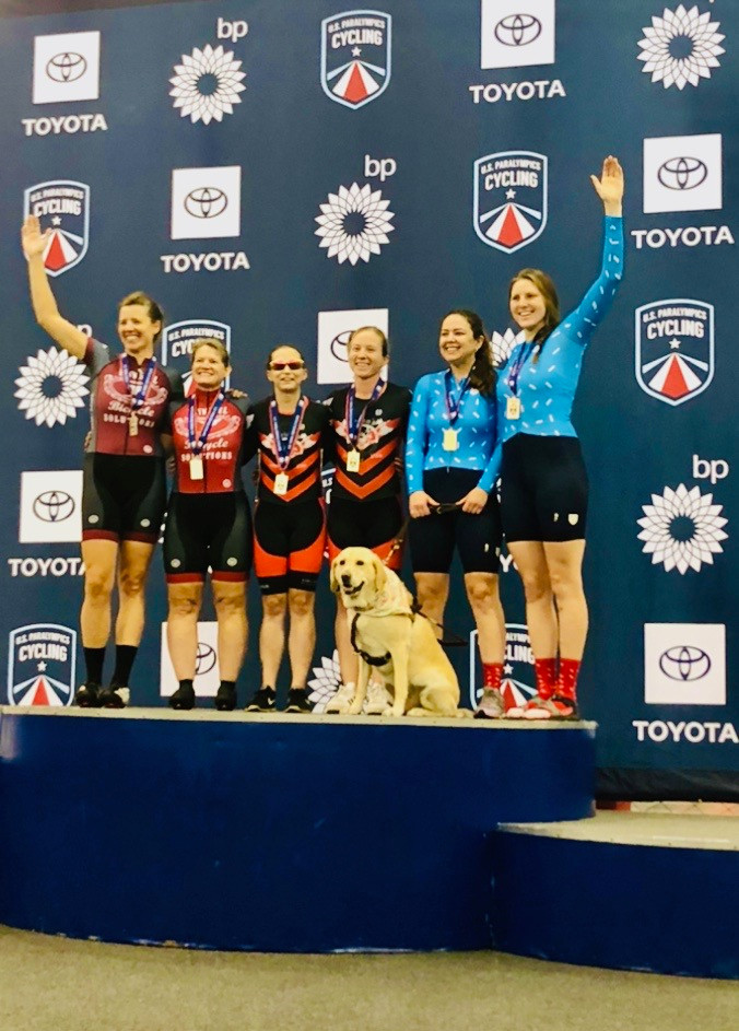PD: Kristin, Zoe and Ash stand on the podium at the U.S. Para Cycling Track Open. Kristin and Zoe are wearing blue cycling suits with black shorts, and Ash has her left arm raised. Four other cyclists are also standing on the podium with them, and everyone has their arms around each other.