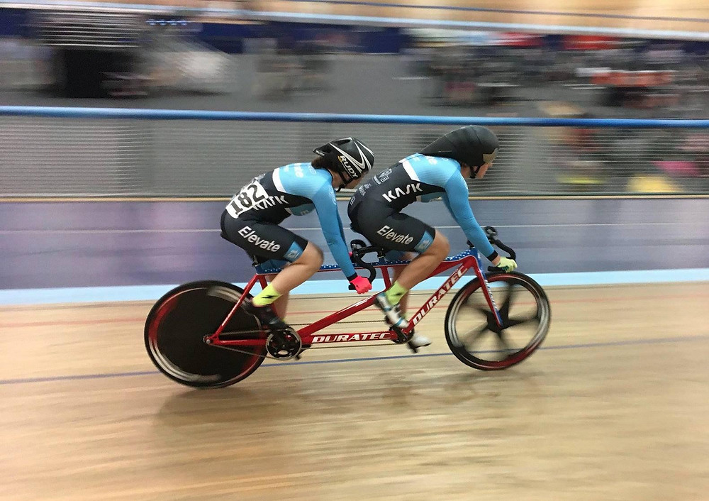 Kristin and her pilot Liesel riding their red Duratec tandem bicycle on the indoor Velodrome Track in Carson, CA. They are both wearing matching light blue cycling kits and black helmets. The background and the bike’s wheels are blurred because the bike is in motion.