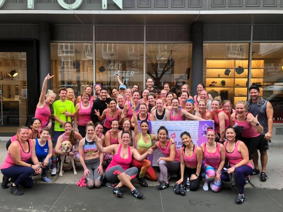 Zoe and Kristin with their cycling friends that are part of the Peloton Monthly Challenge Tribe. They just finished a ride and are outside of the Peloton studio in NYC—this was the first time a service dog had been in the studio. There are about 45 cyclists—mostly pink shirts huddled together showing excitement for finishing their ride. Zoe and Kristin are on the far left. Kristin has her arms draped around Zoe’s neck.