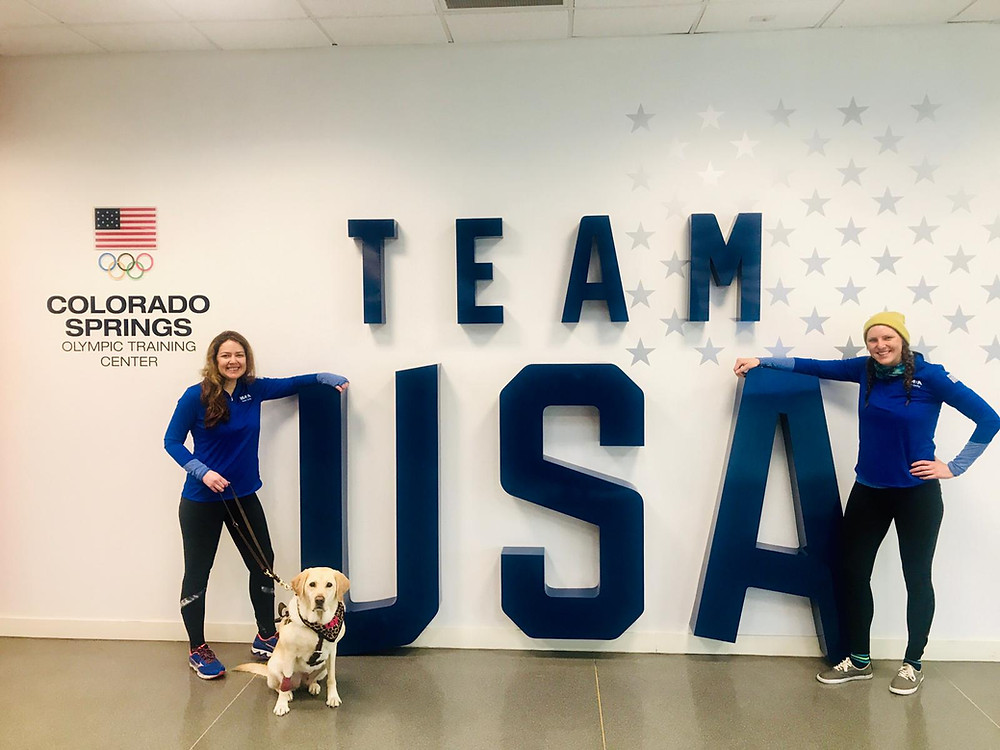 PD: Kristin, Zoe and Ash (Kristin’s Bike Pilot at camp) stand in front of giant blue letters spelling out TEAM USA at the Colorado Spring Olympic Training Center. Kristin and Ash are both smiling, wearing matching blue pullovers. Zoe looks serious and is wearing a leopard print bandana.