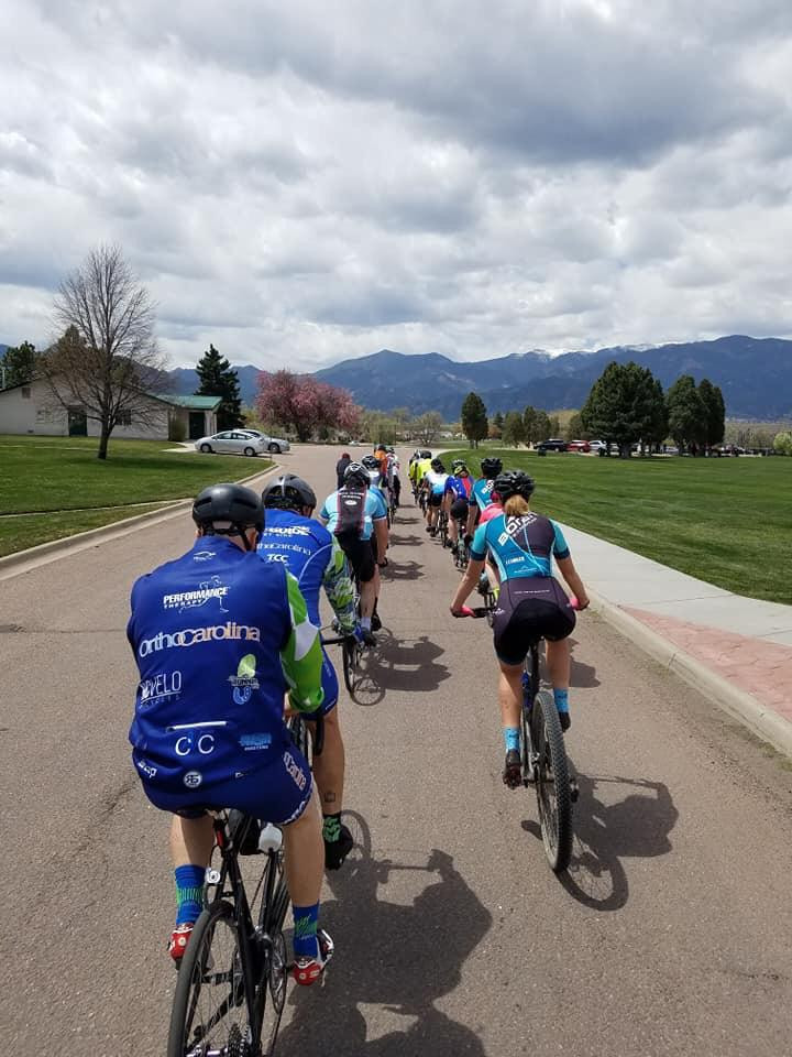 PD: A large group of cyclists are riding in a paceline. We see the cyclists from the back as they ride along the road in two straight lines, side by side.