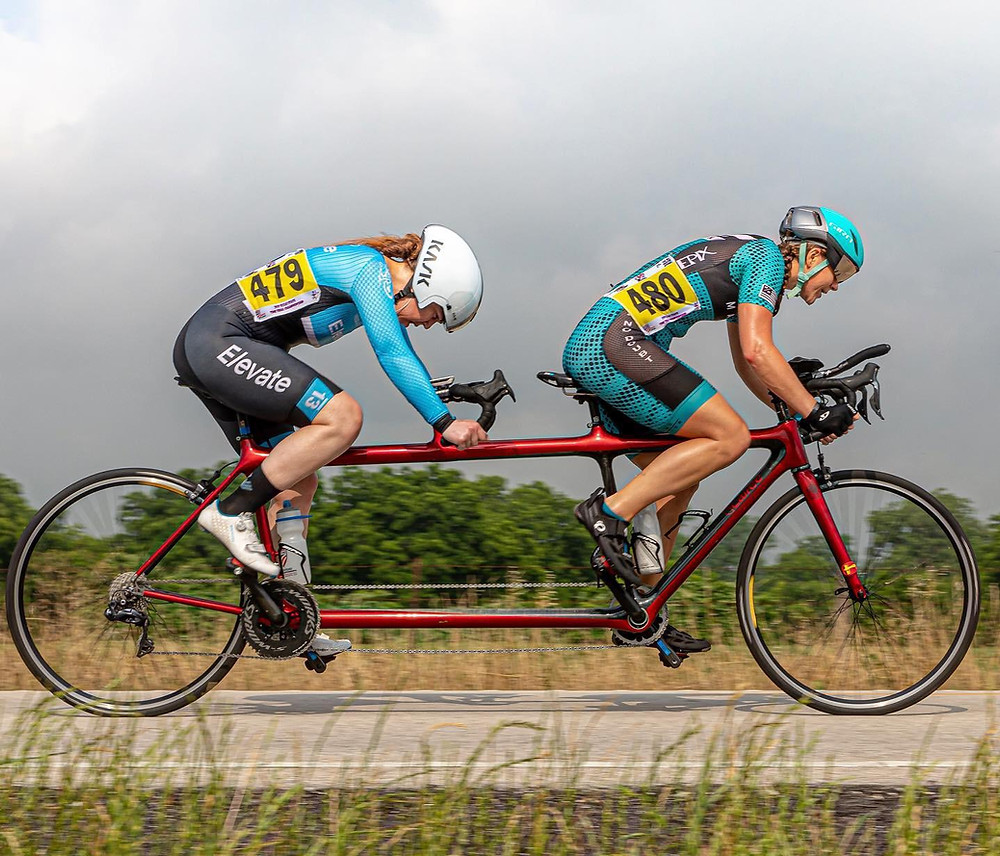 Katie Solbo pilots Kristin Fleschner on a Calfee Design custom candy apple red TT tandem during the Texas State Time Trial 40k. Both Katie and Kristin are in their drops with determined looks on their faces. Katie is wearing a light blue helmet and a light blue and black skinsuit. Kristin is wearing a white helmet and also has on a light blue and black long sleeved skin with the name Elevate going down her right leg. There is long brown grass in the fore and background of the photo and some trees line the grey sky.