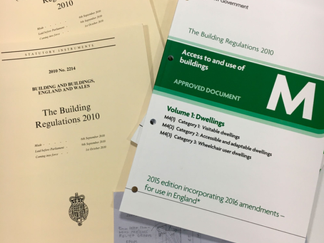 Building regulations documents on a desk include the Building Act 1984. Papers are beige and white with green text accents. Pen nearby.