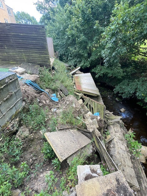 Collapsed fence and retaining wall and debris lie on a muddy slope along a creek. Dense green trees in the background. Earth appears unstable and damaged.