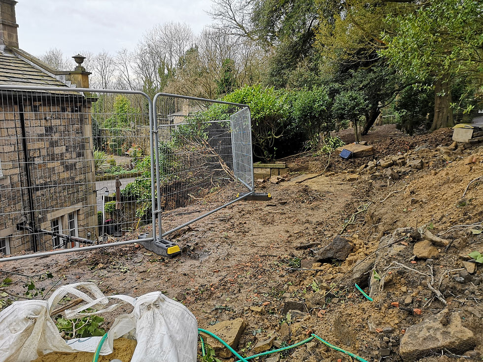 Construction site beside a stone house with metal fencing. Dirt and rocks scattered, surrounded by trees and greenery. Overcast sky.