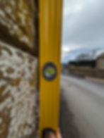 A yellow level against a leaning stone wall shows a green bubble centered. A cloudy sky and empty street are in the background, creating a calm mood.