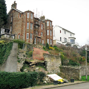 Scaffolded building with red brick and white facade. Collapsed stone retaining wall in foreground. Overcast sky and greenery visible.