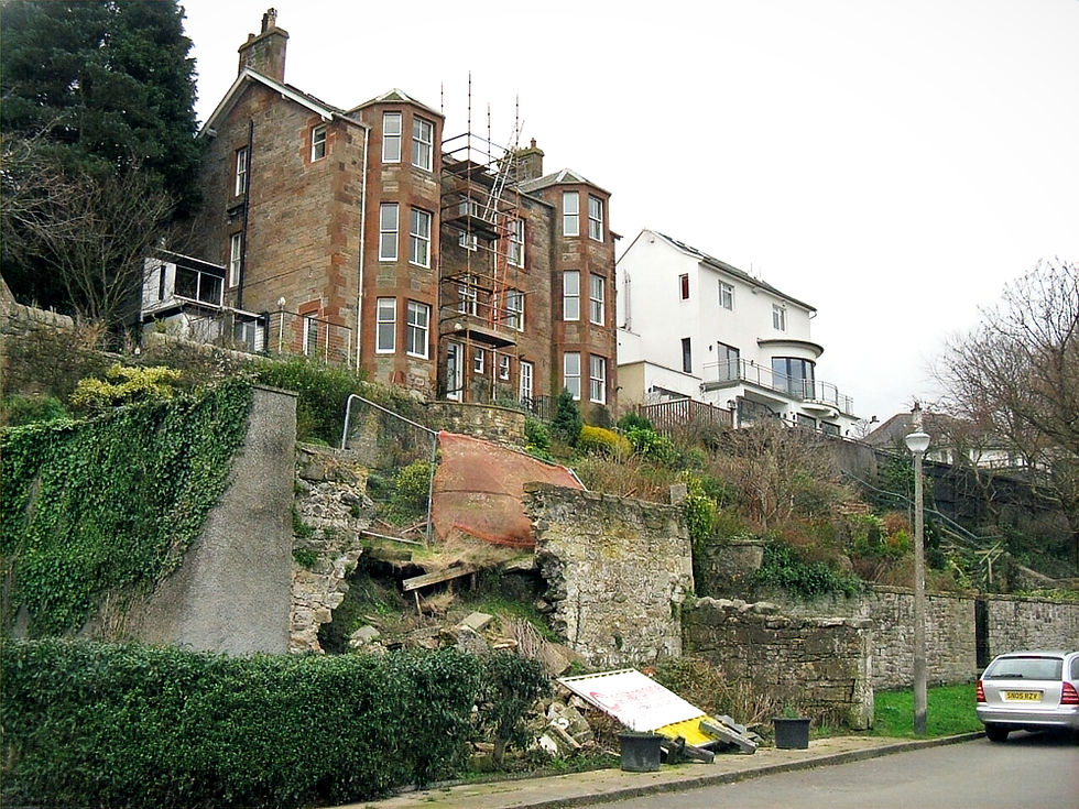 Scaffolded building with red brick and white facade. Collapsed stone retaining wall in foreground. Overcast sky and greenery visible.