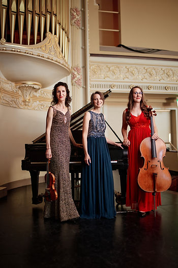 Three musicians in fine dresses stand in front of a grand piano, one of them holds a cello