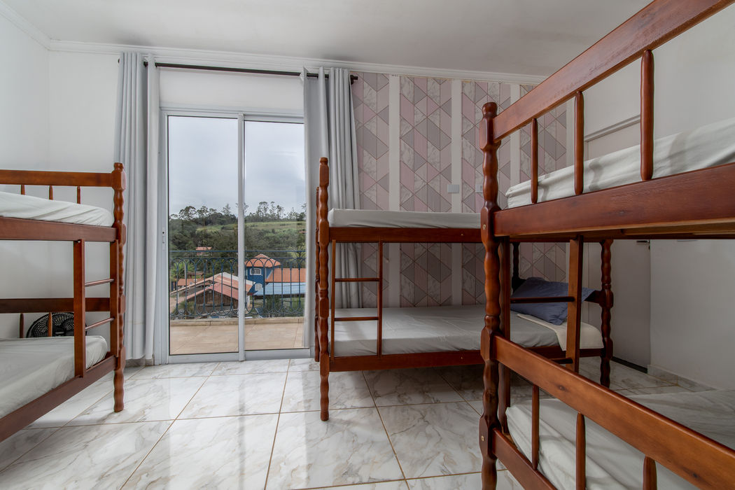 Suíte para seis pessoas com três beliches de madeira, piso de porcelanato marmorizado, papel de parede com estampa geométrica em tons rosados e porta de vidro com sacada abrindo para vista de área verde com construções rurais ao fundo.