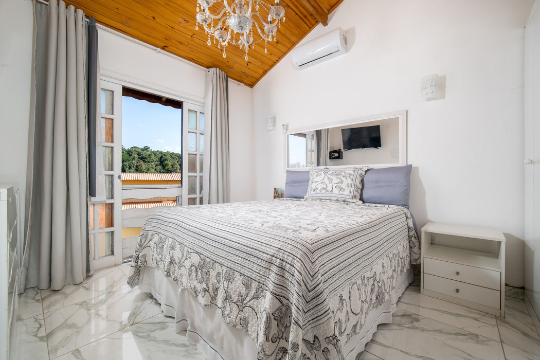 Suíte com cama de casal e roupa de cama em tons de cinza e branco, teto em madeira clara com lustre de cristal, porta-janela com cortinas cinzas e vista para área externa, televisão na parede, mesa de cabeceira branca, ar-condicionado, piso em mármore claro, paredes brancas.
