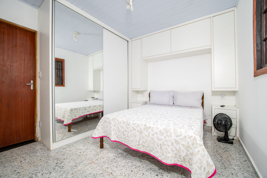 Suíte principal com cama de casal e roupa de cama bege com borda rosa, grande armário branco com porta espelhada à esquerda, armários aéreos e mesas de cabeceira brancas, porta de madeira à esquerda, paredes brancas, teto branco, ventilador, piso em granito claro.