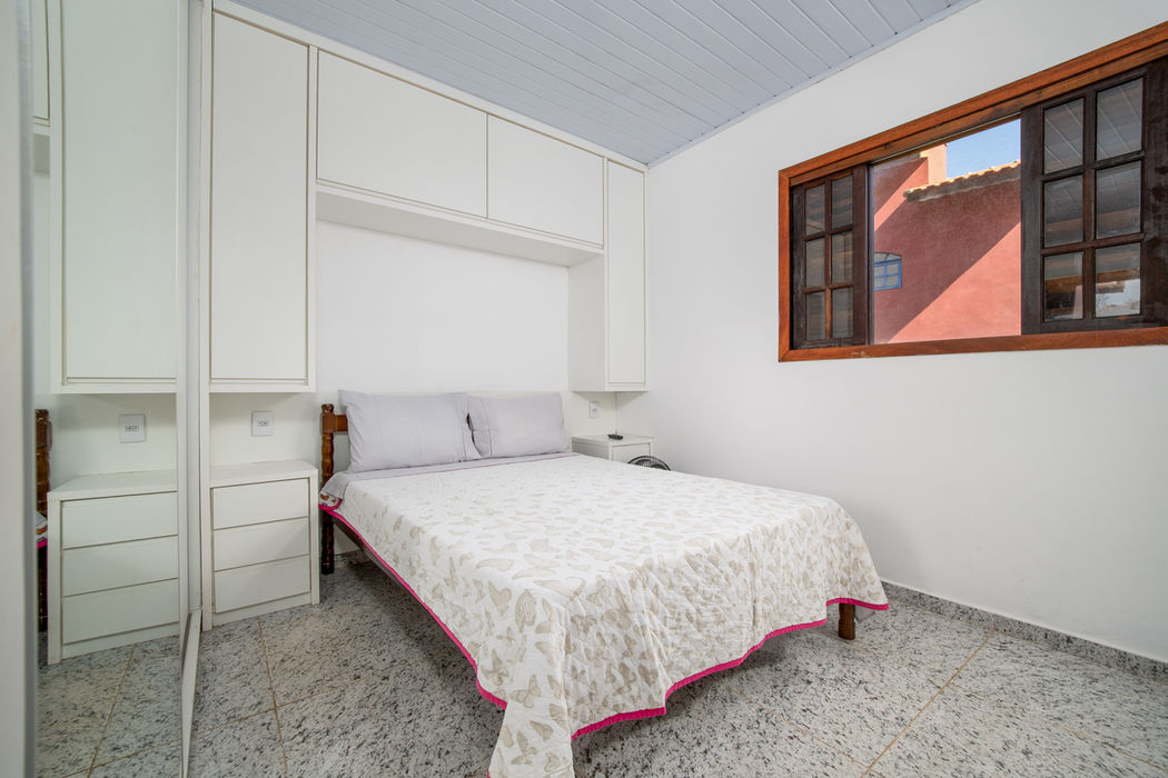 Suíte principal com cama de casal e roupa de cama bege com borda rosa, armários brancos embutidos à esquerda, armários aéreos brancos, janela com moldura de madeira à direita, paredes brancas, teto branco, piso em granito claro.