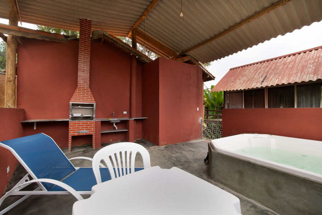 Área de lazer coberta com churrasqueira de tijolos vermelhos, bancadas de apoio, jacuzzi com água à direita, espreguiçadeira azul e mesa com cadeira branca de plástico, paredes em terracota, teto com telhas onduladas, piso cimento queimado, vegetação ao fundo. Jacuzzi ao lado.
