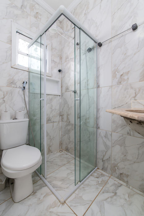 Banheiro da suíte de casal da Casa Branca com vaso sanitário branco à esquerda, box de vidro em L ao centro, janela com moldura branca e armário branco no box, revestimento em mármore bege com veios, bancada em mármore à direita, teto branco, piso em mármore.