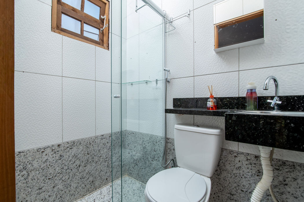 anheiro da suíte com box de vidro à esquerda, vaso sanitário branco ao centro, pia com bancada em granito preto à direita, revestimento em cerâmica branca na parte superior e granito claro na base, janelas com moldura de madeira, espelho na parede, piso em granito claro.