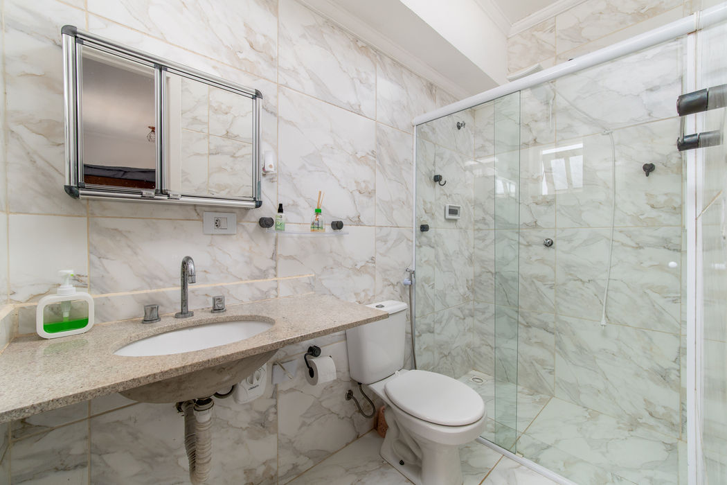 Banheiro revestido com porcelanato marmorizado branco, bancada de granito com cuba embutida e torneira cromada, espelho com moldura de alumínio, vaso sanitário e box de vidro temperado com chuveiro elétrico.