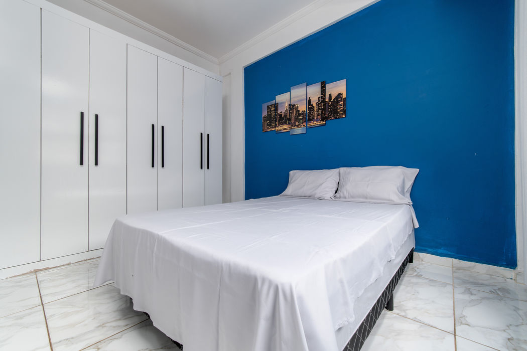 Suíte de casal da Casa Branca com cama de casal e roupa de cama branca, parede azul atrás da cama com quadro decorativo de cidade, grande armário branco à esquerda, teto branco, piso em mármore claro.