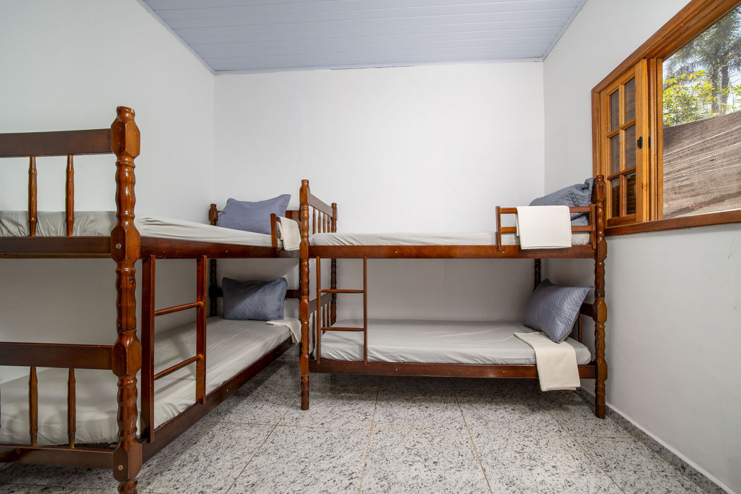 Suíte para quatro pessoas com dois beliches de madeira, roupa de cama branca com travesseiros cinza, paredes brancas, teto branco, janela com moldura de madeira à direita, piso em granito claro.