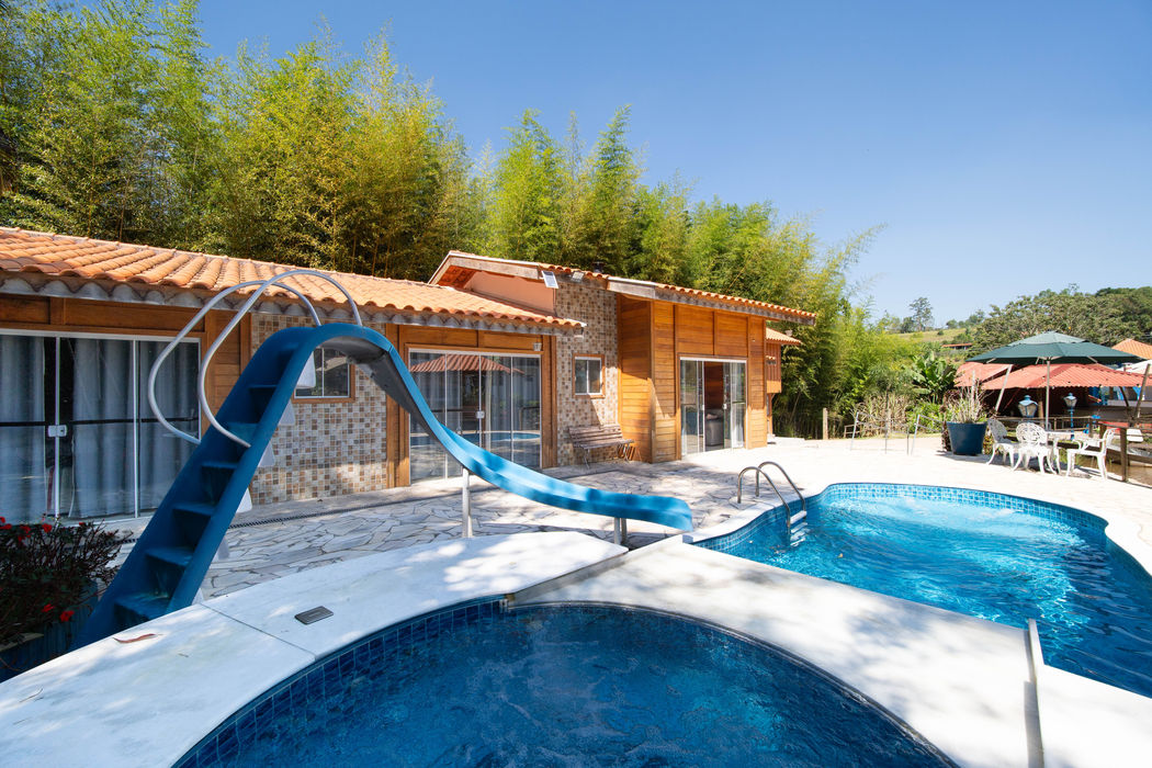 Área de lazer da Casa do Lago com piscina, escorregador azul, deck em pedra clara, casa com paredes em madeira e tijolos, telhado com telhas laranjas, amplas janelas de vidro, vegetação densa ao fundo, guarda-sóis e móveis de jardim, céu azul.