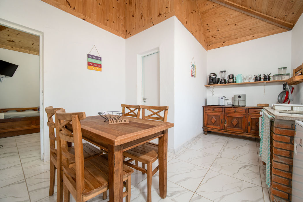 Cozinha e sala de jantar integrada com mesa quadrada de madeira e 4 cadeiras, teto em madeira clara, paredes brancas, buffet de madeira escura com eletrodomésticos, prateleira de madeira na parede, placas decorativas, piso em cerâmica clara, porta branca ao fundo.