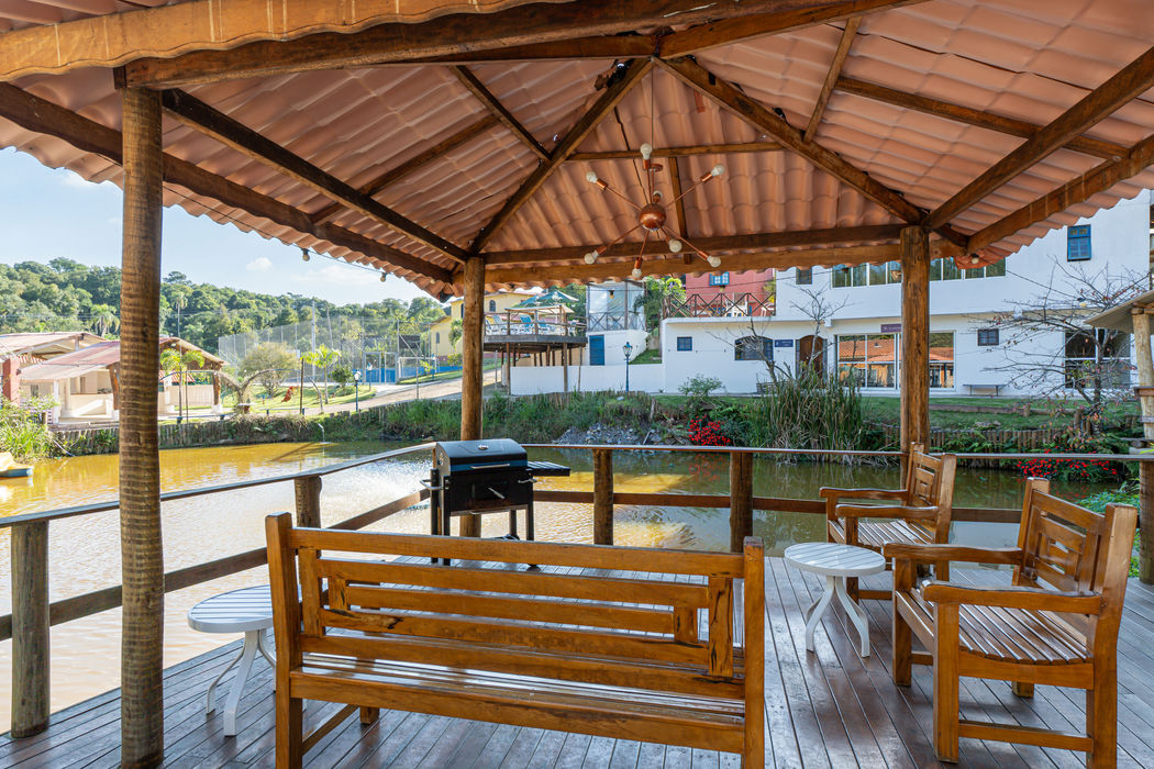 Deck coberto com churrasqueira, banco de madeira, mesa e cadeiras de madeira, teto com telhas e estrutura de madeira aparente, piso em madeira, vista para lago e propriedade com casas e vegetação ao fundo, lustre no teto.
