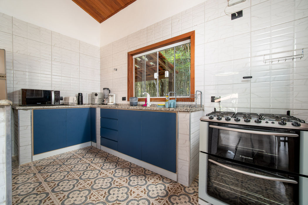 Cozinha com armários inferiores azuis e bancada em granito bege, paredes revestidas em azulejo branco, fogão inox à direita, microondas e eletrodomésticos sobre a bancada, janela com moldura de madeira ao centro com vista para o quintal, teto branco com detalhe em madeira, piso com ladrilho hidráulico decorativo.