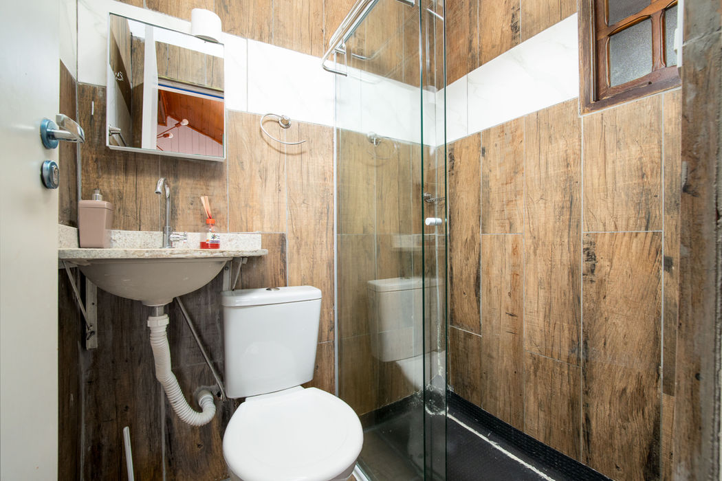 Banheiro da suíte com pia suspensa com bancada em granito à esquerda, vaso sanitário branco, box de vidro à direita, revestimento em porcelanato que imita madeira, armário espelhado acima da pia, teto em madeira, janela com moldura de madeira, paredes brancas na parte superior, piso escuro.