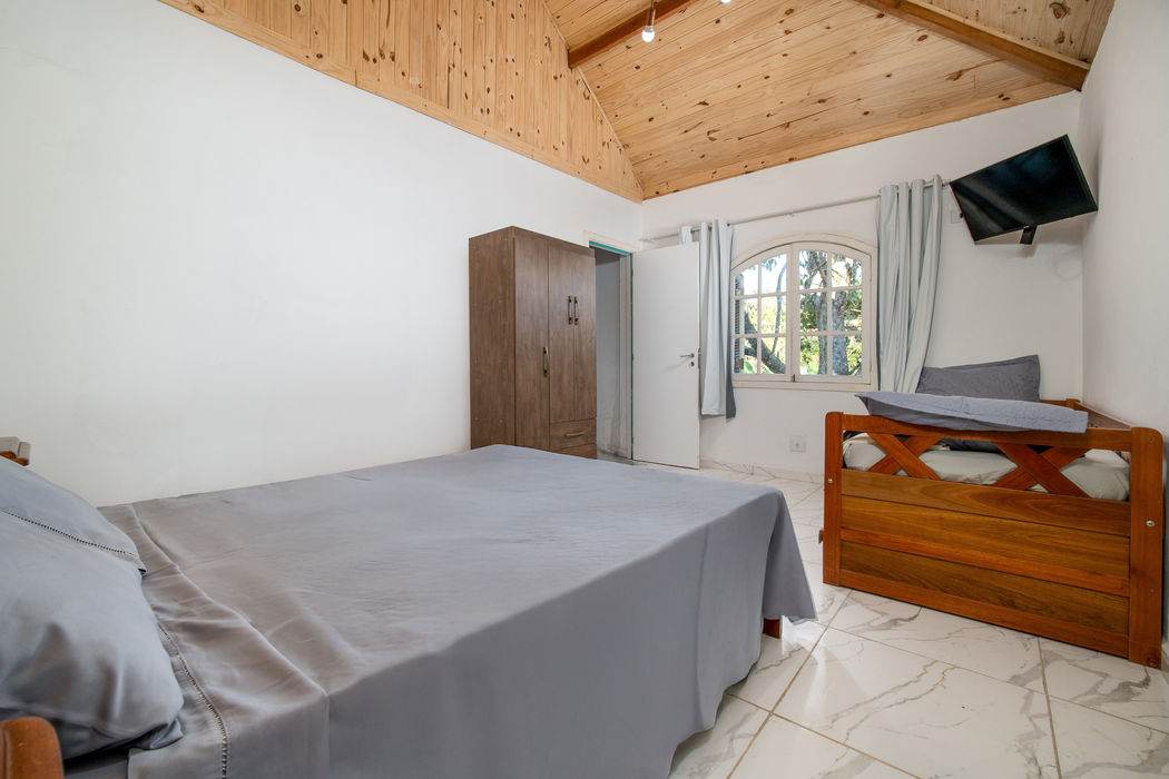 Cama de solteiro com roupa de cama cinza, teto em madeira clara, armário de madeira escura ao fundo, janela em arco com moldura branca e cortina, porta branca, televisão na parede, paredes brancas, piso em cerâmica clara.