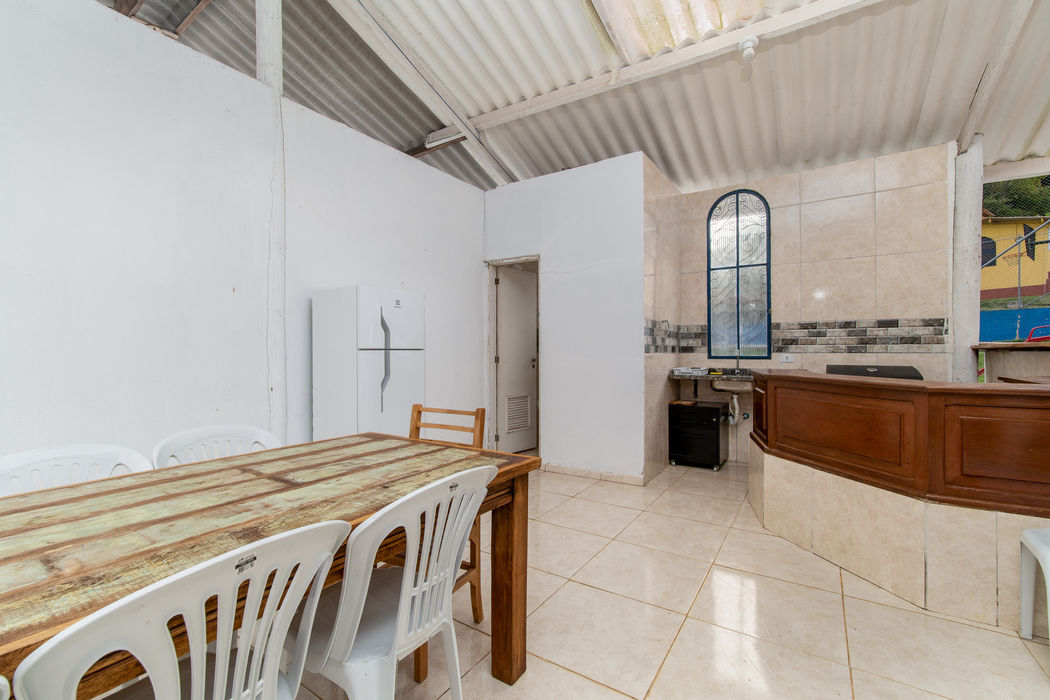 Interior da área de churrasqueira com mesa rústica de madeira demolição e cadeiras plásticas brancas, balcão curvo de madeira, geladeira branca, pia com revestimento em pedra, janela em arco com esquadria azul, piso de porcelanato bege.