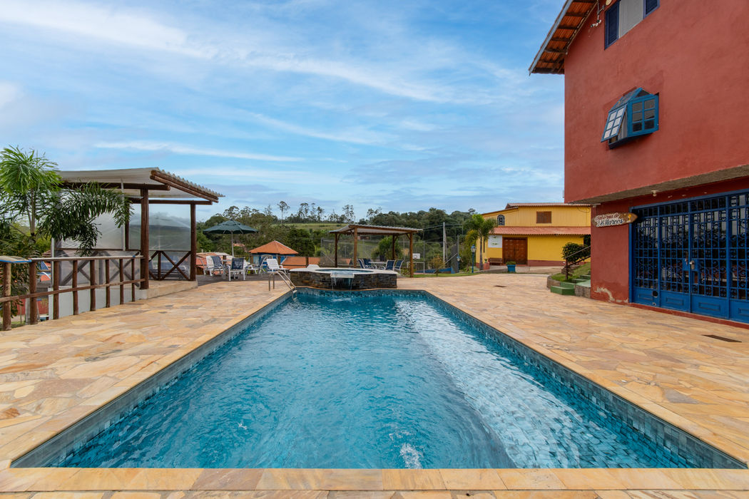 Área de piscina com piso em pedra bege, piscina retangular com água azul cristalina, deck de madeira com espreguiçadeiras e guarda-sol.