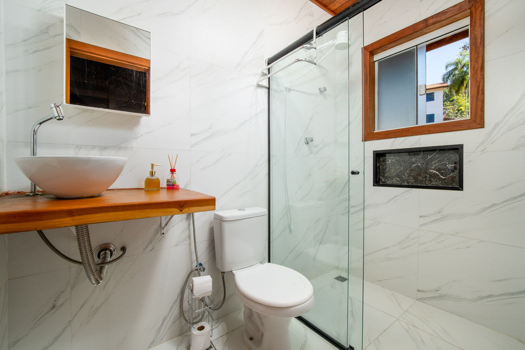 Banheiro da suíte com pia de apoio branca sobre bancada de madeira suspensa à esquerda, vaso sanitário branco ao centro, box de vidro à direita, revestimento em porcelanato branco com efeito mármore, espelho com moldura de madeira sobre a pia, janela com moldura de madeira, nicho preto embutido na parede, teto branco com detalhe em madeira, piso em porcelanato branco.
