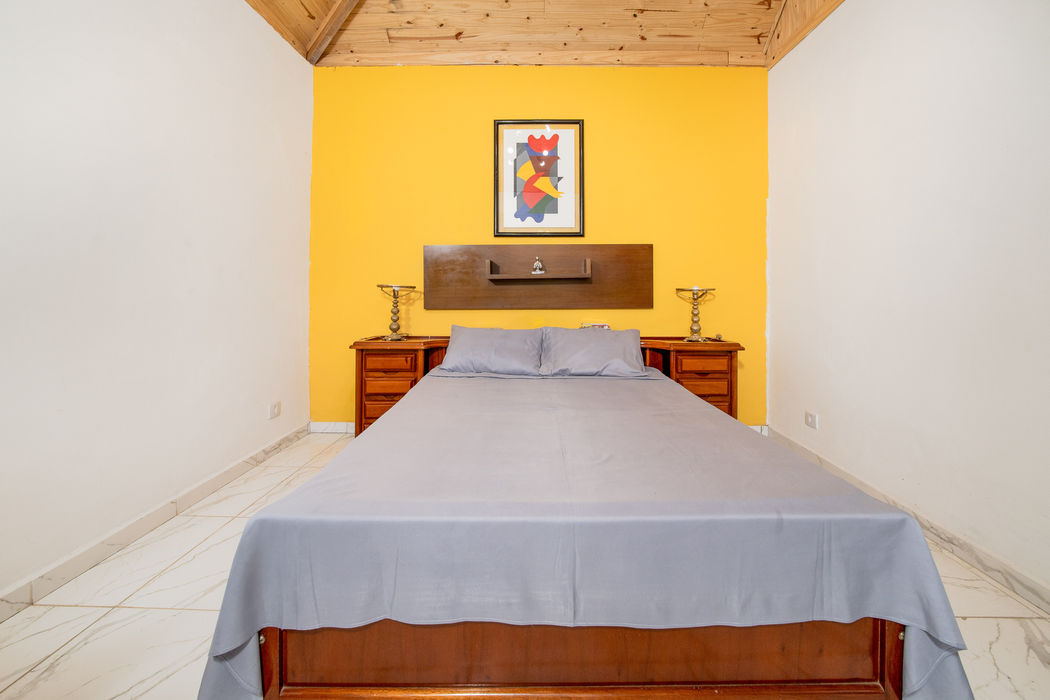 Quarto com cama de casal de madeira e roupa de cama cinza, parede amarela ao fundo com quadro colorido abstrato, cabeceira de madeira suspensa, mesas de cabeceira de madeira com luminárias, teto em madeira clara, paredes brancas, piso em cerâmica clara.