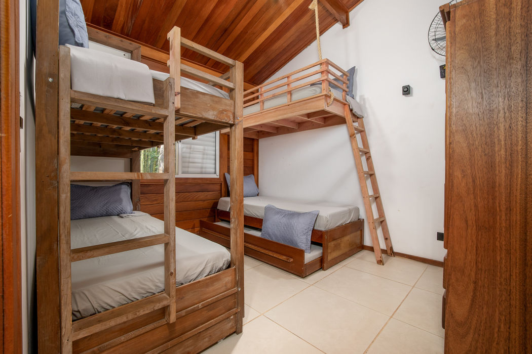 Quarto para cinco pessoas com dois beliches de madeira e cama auxiliar, roupa de cama branca com travesseiros cinza, teto em madeira, paredes brancas, janela, escada de madeira para acesso aos beliches superiores, armário de madeira à direita, ventilador na parede, piso em cerâmica clara.