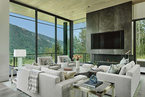 Large window view into the dining room of Shadow Mountain Residence in Aspen, showing contemporary interiors with natural textures, stone walls, and a connection to surrounding evergreens.