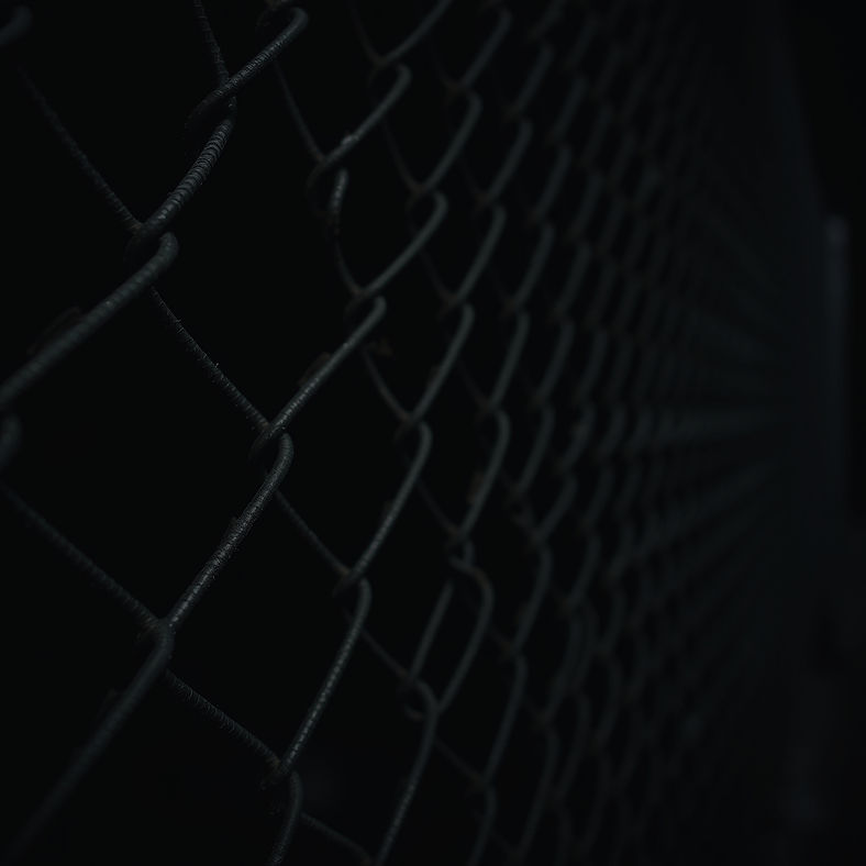 A gritty, dark background featuring a close-up view of a weathered chainlink fence. The fe