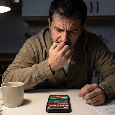Homem preocupado olhando para o celular com aplicativo de apostas online aberto na mesa, representando o estresse financeiro e a ansiedade do vício em jogos.