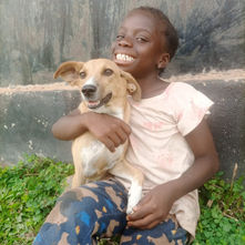 A girl sitting on grass with a concrete wall behind them and she is holding a tan and white dog in her lap both of them smiling in Liberia.