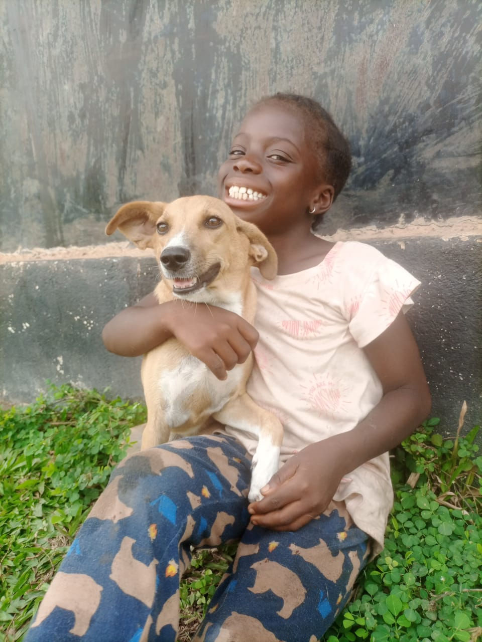 A girl sitting on grass with a concrete wall behind them and she is holding a tan and white dog in her lap both of them smiling in Liberia.