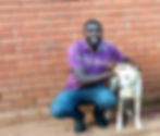 Man in purple shirt smiling next to light-colored dog against red brick wall at the animal shelter in Uganda.